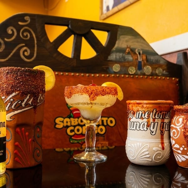 A Corona beer bottle and three festive, chili-rimmed drinks with lemon slices on a dark table.