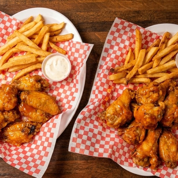 Two plates of chicken wings and french fries with dipping sauce on red and white checkered paper.